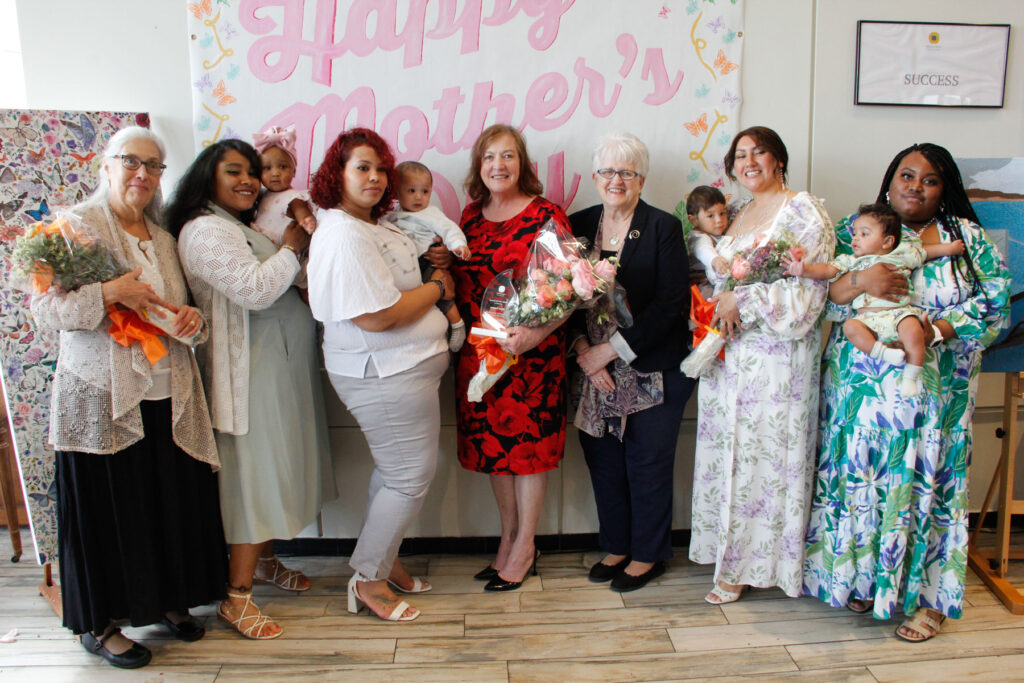 A group of women, including mothers in recovery, holding babies and bouquets standing in front of a Happy Mother's Day banner